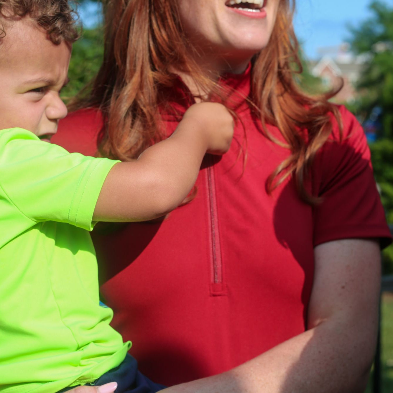 Detail shot of woman wearing red Betty Quarter Zip Dress while holding son at the playground with friends showcasing the dress's versatility, durability, and 4-way stretch performance fabric.