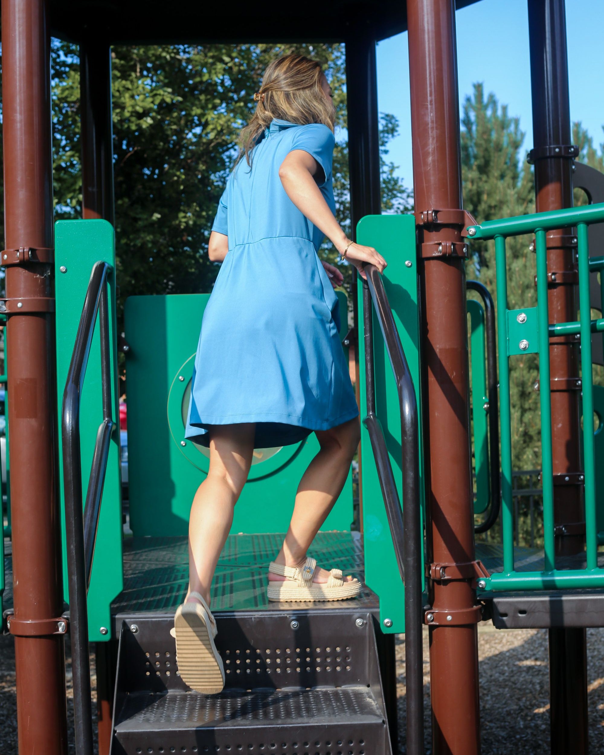 Person in a blue Clarece Polo Dress climbing a playground structure with trees in the background