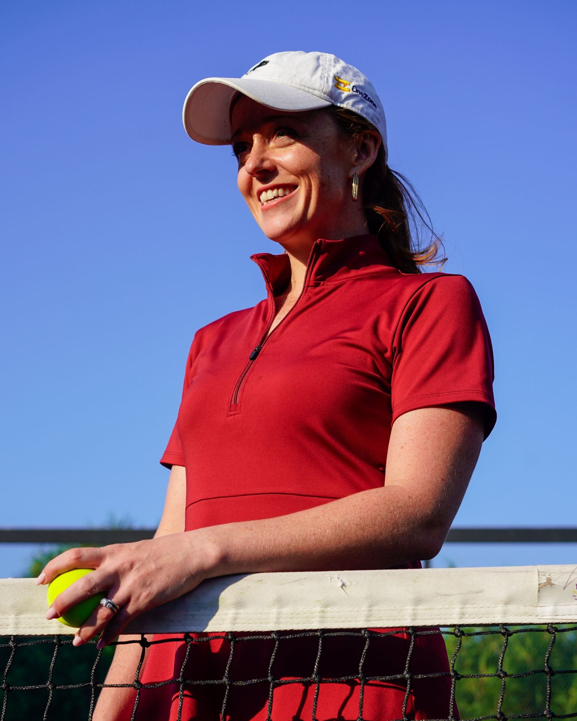 Detail shot of woman smiling while wearing red Betty Quarter Zip Dress at the tennis court, featuring the high-quality performance fabrics and functional zipper in the neckline.