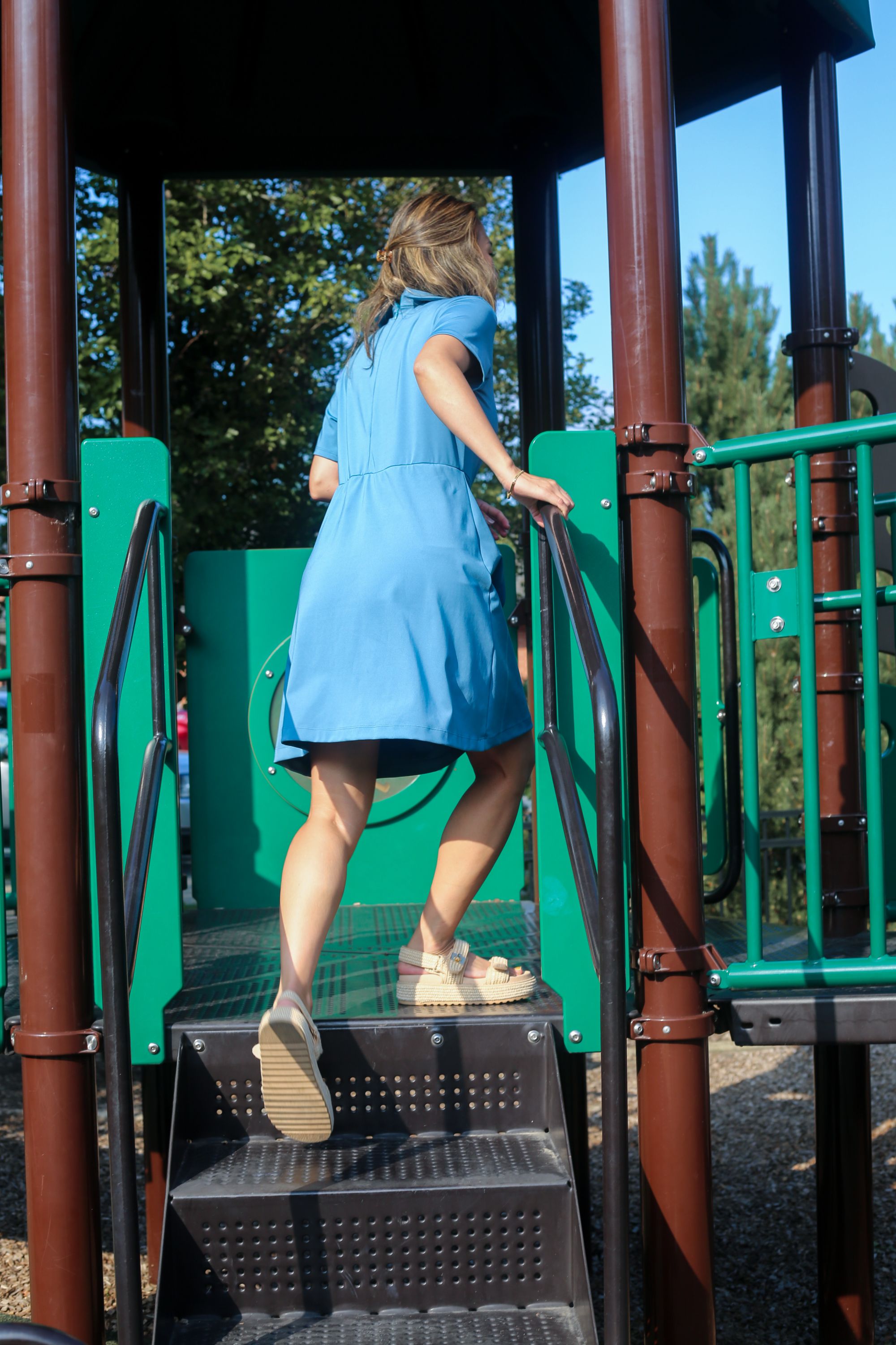 Person in a blue polo-collared dress climbing a playground structure with trees in the background