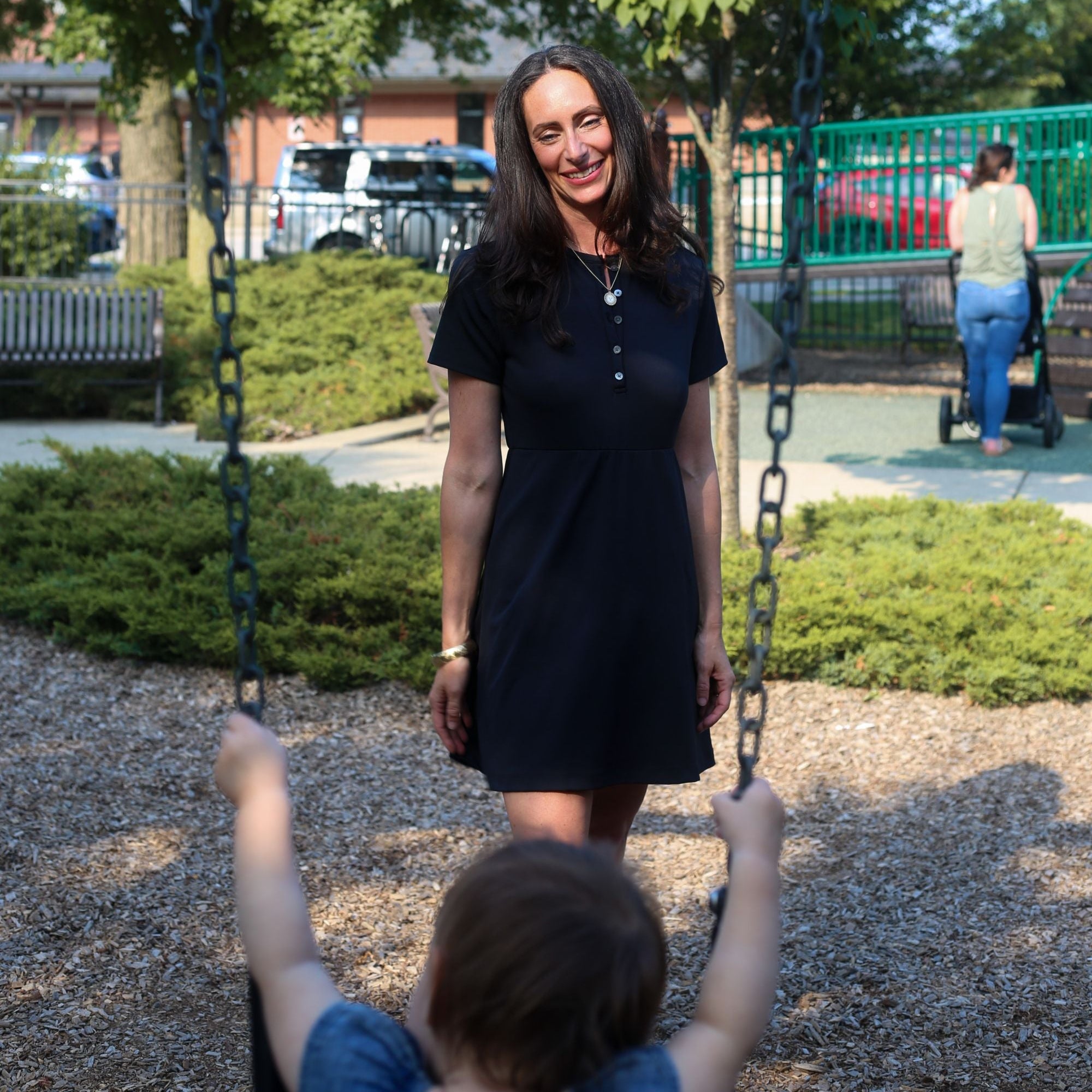 Woman wearing Ace Henley Dress in black at the park swings showcasing the dress's UV protection, moisture-wicking, and versatility. 