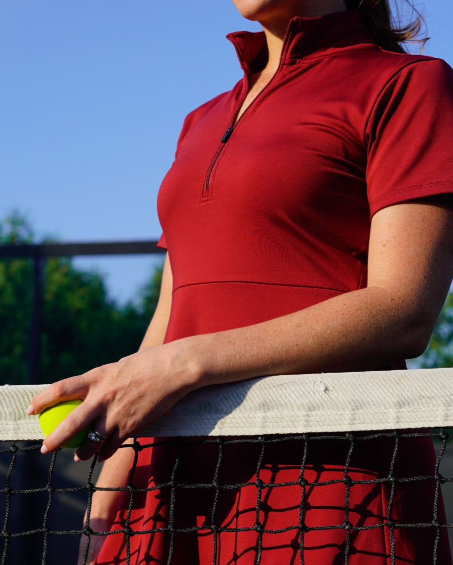 Detail shot of woman wearing red Betty Quarter Zip Dress at the tennis court, featuring the high-quality performance fabrics and functional zipper in the neckline.